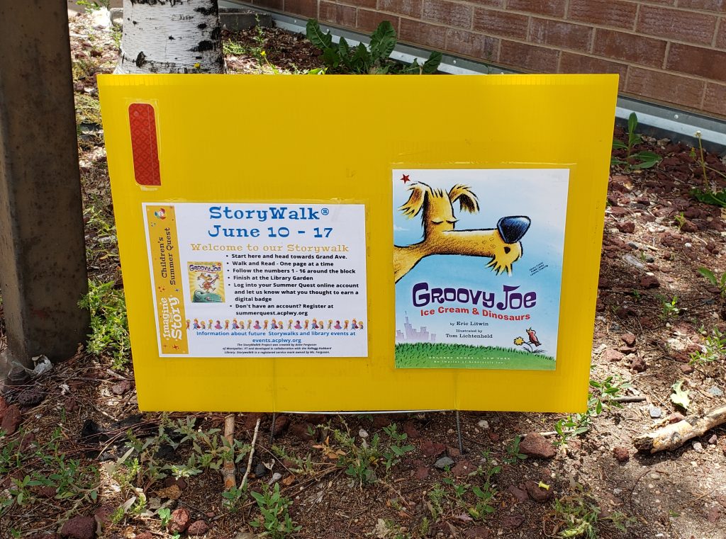 An image showing a yellow sign outside that has the front of the picture book titled Groovy Joe: Ice Cream and Dinosaurs and instructions for completing a Story Walk (R): June 10-17