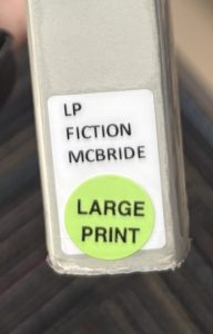 A closeup of the spine on the book "The Heaven & Earth Grocery Store," focusing on the spine label. The spine label reads "LP FICTION MCBRIDE" and features a green circle sticker that reads "LARGE PRINT."
