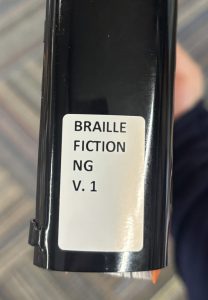 A closeup of the spine on the Braille book "Little Fires Everywhere," focusing on the spine label. The spine label reads "BRAILLE FICTION NG V.1."